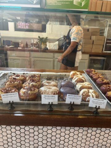 A display of various pastries, including Butterscotch Bacon, Peaches & Cream, Boston Cream Puffs, and Fluffernutter, with a person in the background.