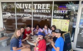 Group of people sitting at an outdoor table in front of a Mediterranean grille restaurant called The Olive Tree. They are eating and smiling at the camera.