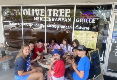 Group of people sitting at an outdoor table in front of a Mediterranean grille restaurant called The Olive Tree. They are eating and smiling at the camera.