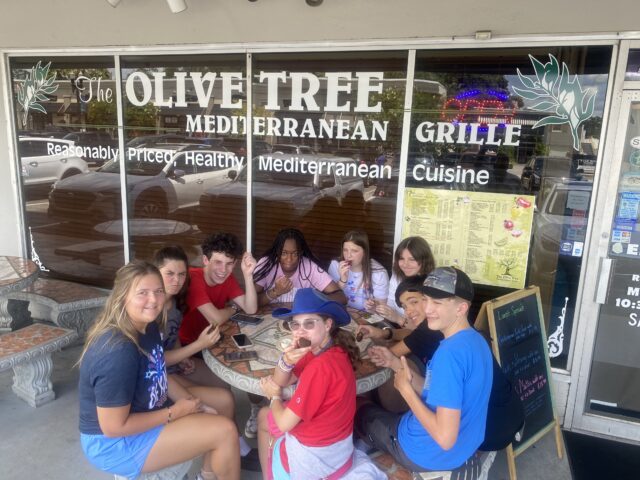 Group of people sitting at an outdoor table in front of a Mediterranean grille restaurant called The Olive Tree. They are eating and smiling at the camera.