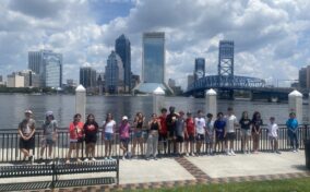 A group of people stands by a river with a city skyline and bridge in the background. It's a sunny day with some clouds.