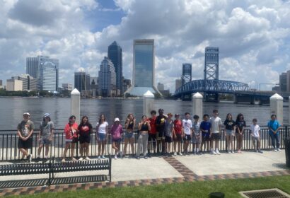 A group of people stands by a river with a city skyline and bridge in the background. It's a sunny day with some clouds.