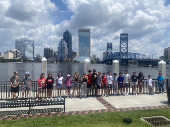 A group of people stands by a river with a city skyline and bridge in the background. It's a sunny day with some clouds.