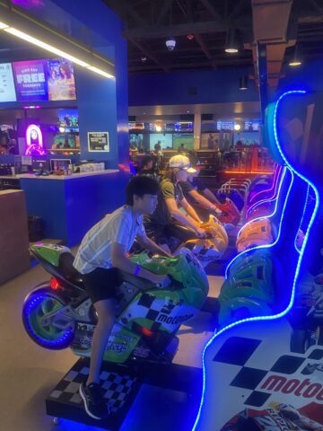 People are engaged in a motorcycle racing arcade game in a dimly lit entertainment venue.