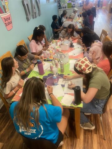 A group of people are gathered around a table, engaged in a craft activity with paints and canvases in a room with wooden floors and light blue walls.