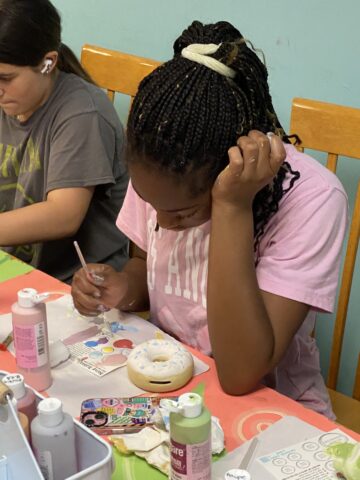 A girl in a pink shirt paints a circular object at a table. She has braided hair tied up. Art supplies and paint bottles are on the table. Another person is seated beside her.