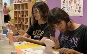 Two young women are painting ceramic figures at a table in a colorful studio. Art supplies and paint bottles are visible around them.