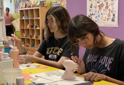 Two young women are painting ceramic figures at a table in a colorful studio. Art supplies and paint bottles are visible around them.