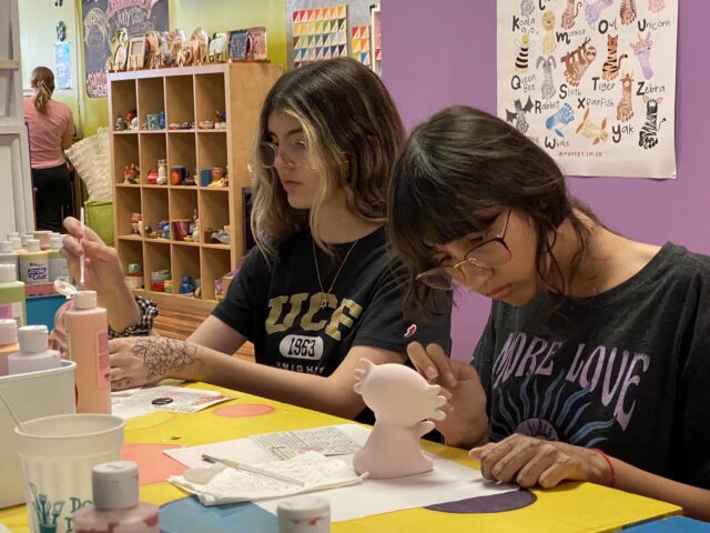 Two young women are painting ceramic figures at a table in a colorful studio. Art supplies and paint bottles are visible around them.