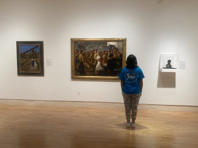 Person in a blue shirt observes paintings and a sculpture in an art gallery.