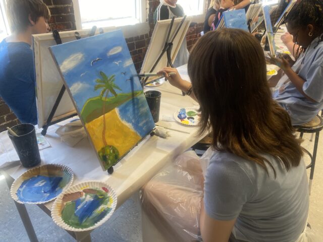 People are painting ocean scenes on canvases in a classroom setting.