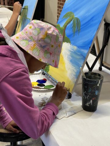 Child in a pink hat painting a beach scene on a canvas at an art class, with paint supplies on the table.