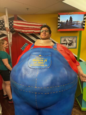 A person stands inside a large, blue, novelty costume at Ripley's Believe It or Not! exhibit, with a U.S. flag in the background and a mounted screen displaying an image.