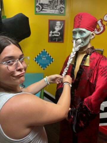 A person playfully grabs a long braid of a wax figure dressed in red and black, in a room with yellow walls and vintage decor.