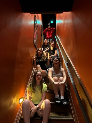 Six people sitting on stairs with orange walls, posing for a group photo. The person at the top appears to be making a gesture with their arms.