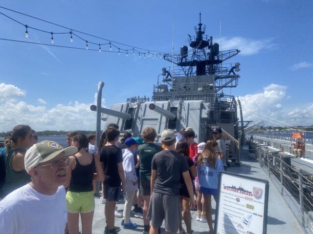 A group of people stands on the deck of a naval ship, observing the surroundings. The sky is clear and sunny.