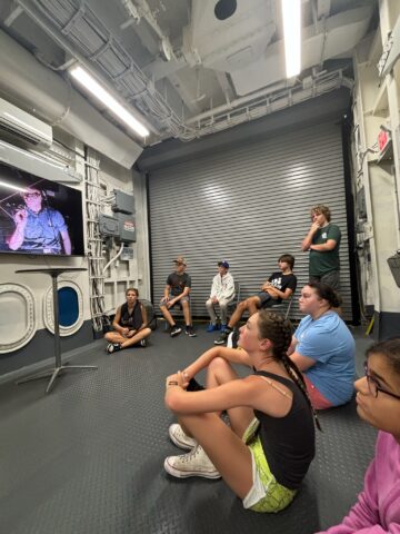 A group of people sit attentively on the floor of a room with metal walls, watching a presentation on a television screen.