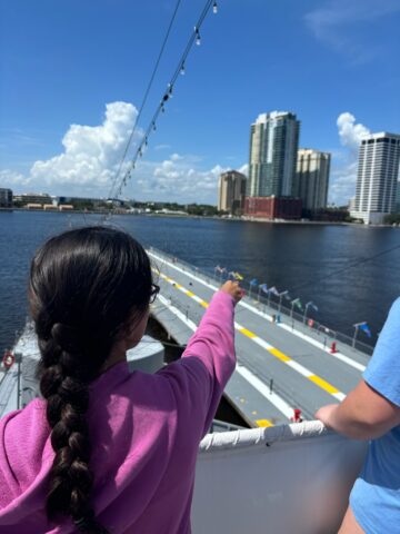 Person in a pink hoodie and braid pointing toward a cityscape from a ship deck, with tall buildings and a body of water under a clear blue sky.