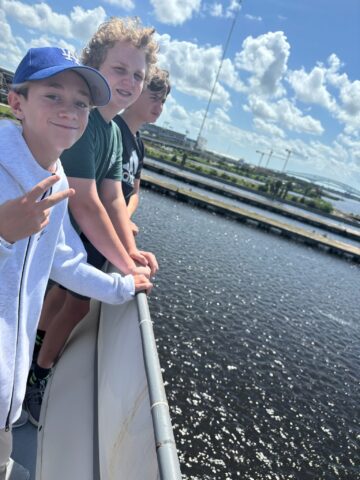 Four people stand on a boat deck, overlooking the water on a sunny day. One person in the foreground wears a blue hat and makes a peace sign.