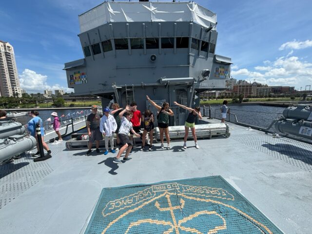 A group of people posing on the deck of a ship. Some are doing the dab dance move. The city skyline and blue sky are visible in the background.