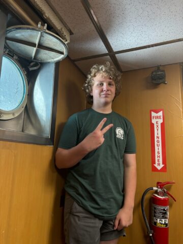 Teen boy in a green T-shirt making a peace sign while standing next to a fire extinguisher inside a wooden-paneled room with a round window.