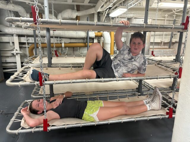 Two people relax on metal-framed bunk beds in an industrial-looking room, with overhead pipes visible.