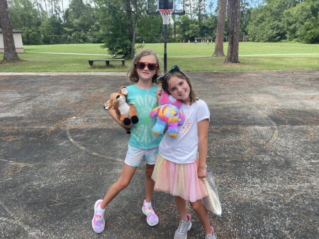 Two children stand outdoors on a basketball court. One holds a stuffed animal, and the other has a unicorn plush. Trees and grass are visible in the background.