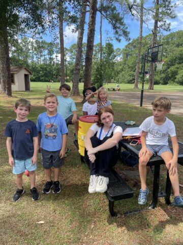 A group of children sitting and standing around a picnic table outdoors, with trees and a basketball court in the background.