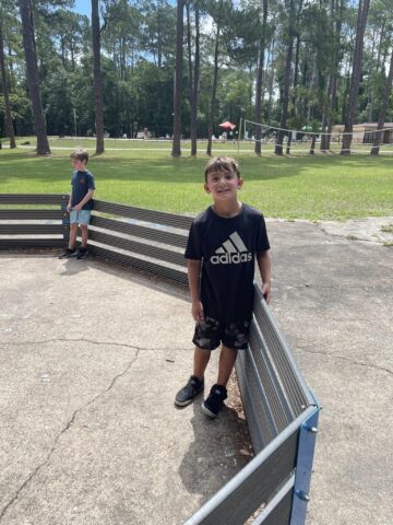 Two boys stand in a fenced circular area outdoors, with one boy facing the camera and the other looking away. Trees and a playground are in the background.