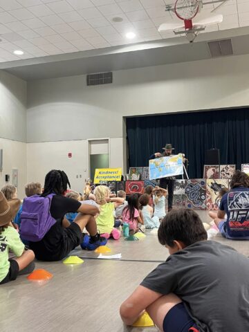 A man on stage shows a world map to seated children in a school gym. A sign reads "Kindness and Acceptance.