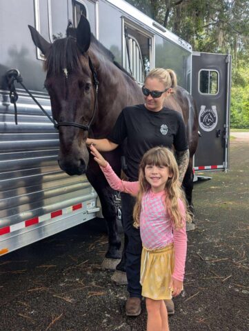 A woman and a child stand next to a dark horse, which is tethered to a trailer. The child is gently petting the horse's nose.