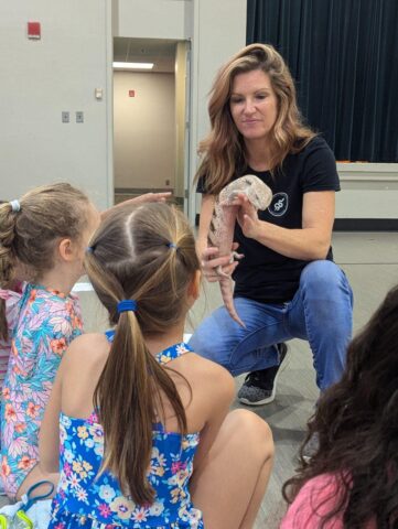 A woman kneels and holds a lizard in front of a group of seated children in a classroom.