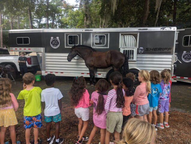 A group of children observes a large black horse standing outside a sheriff's trailer in a wooded area.