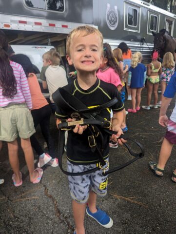 A smiling young boy holds horse tack while standing in front of a group of children near a horse trailer.