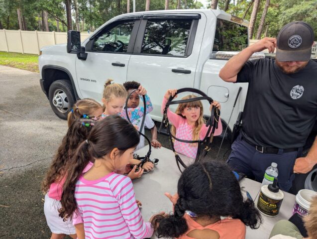 Children gather around a table with horse harnesses and a man by a white truck.