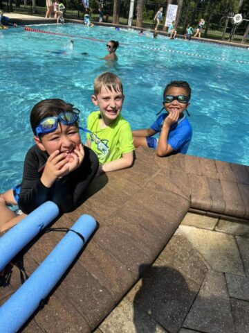 Three children in swimwear are sitting by the edge of a pool on a sunny day, with pool noodles visible in the foreground.