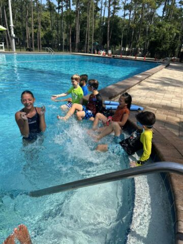 Children sitting by a pool laughing and splashing water under a clear sky, surrounded by trees.