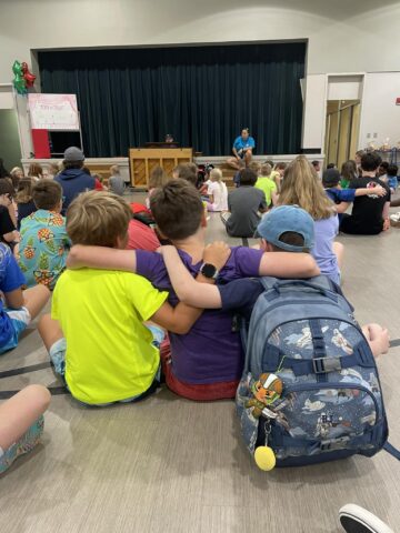 Children sitting on the floor in a gymnasium, facing a stage. Three friends in colorful shirts sit in the front with arms around each other, one wearing a backpack. A speaker stands on the stage.