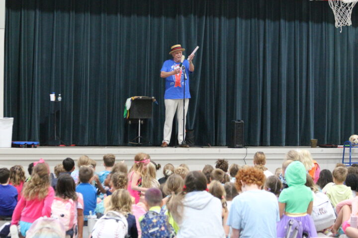 A man on stage performs with a magic wand in front of a seated audience of children.