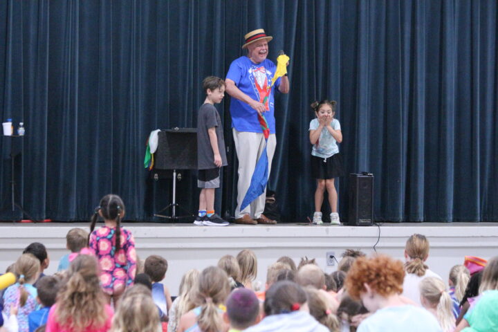 A performer entertains a group of children on stage, holding a colorful prop, with two kids beside him. The audience of kids watches from the floor in front of the stage.