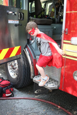A young boy wearing a superhero cape on a fire truck.