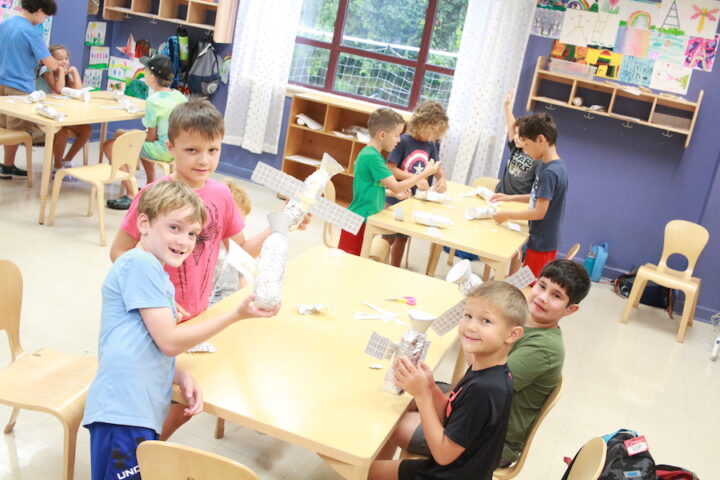 A group of children are sitting at a table making paper airplanes.