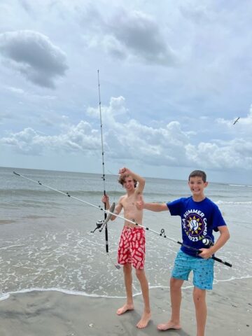 Two boys standing on the beach holding fishing rods.