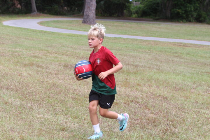 A young boy in a red and green jersey runs on grass while holding a blue and red ball.