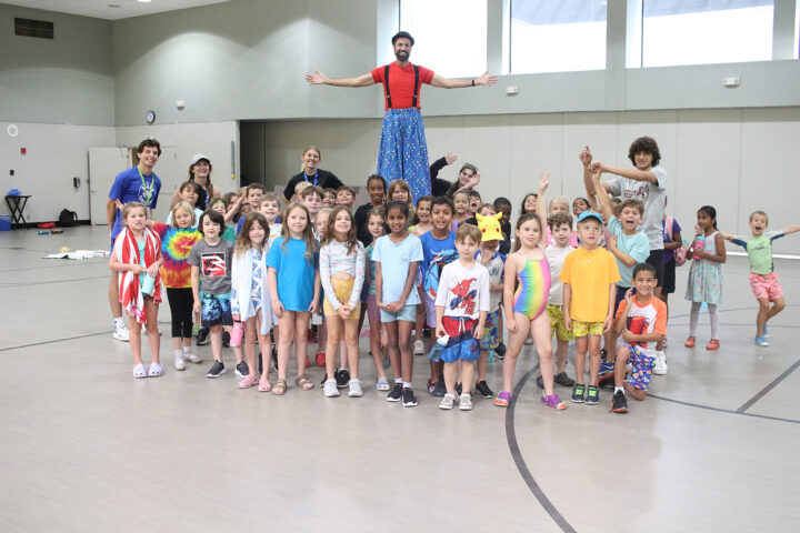 A group of children and adults pose in a gymnasium, with an entertainer on stilts at the back. Everyone is smiling and some are gesturing playfully.