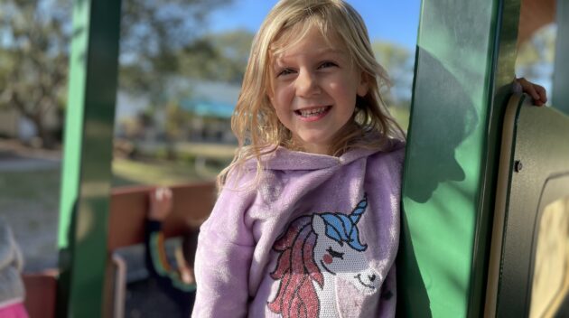 A little girl wearing a unicorn sweatshirt on a playground.