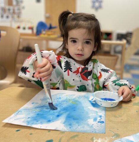 A little girl with a paint brush at a table.