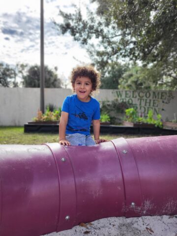 A young boy sitting on a purple pipe in a playground.