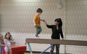 A young gymnast walks on the balance beam with the gymnastics instructors help