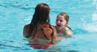 Two people are in a swimming pool, facing each other. One is a child sticking out their tongue, and the other is an adult with wet hair wearing a red swimsuit.